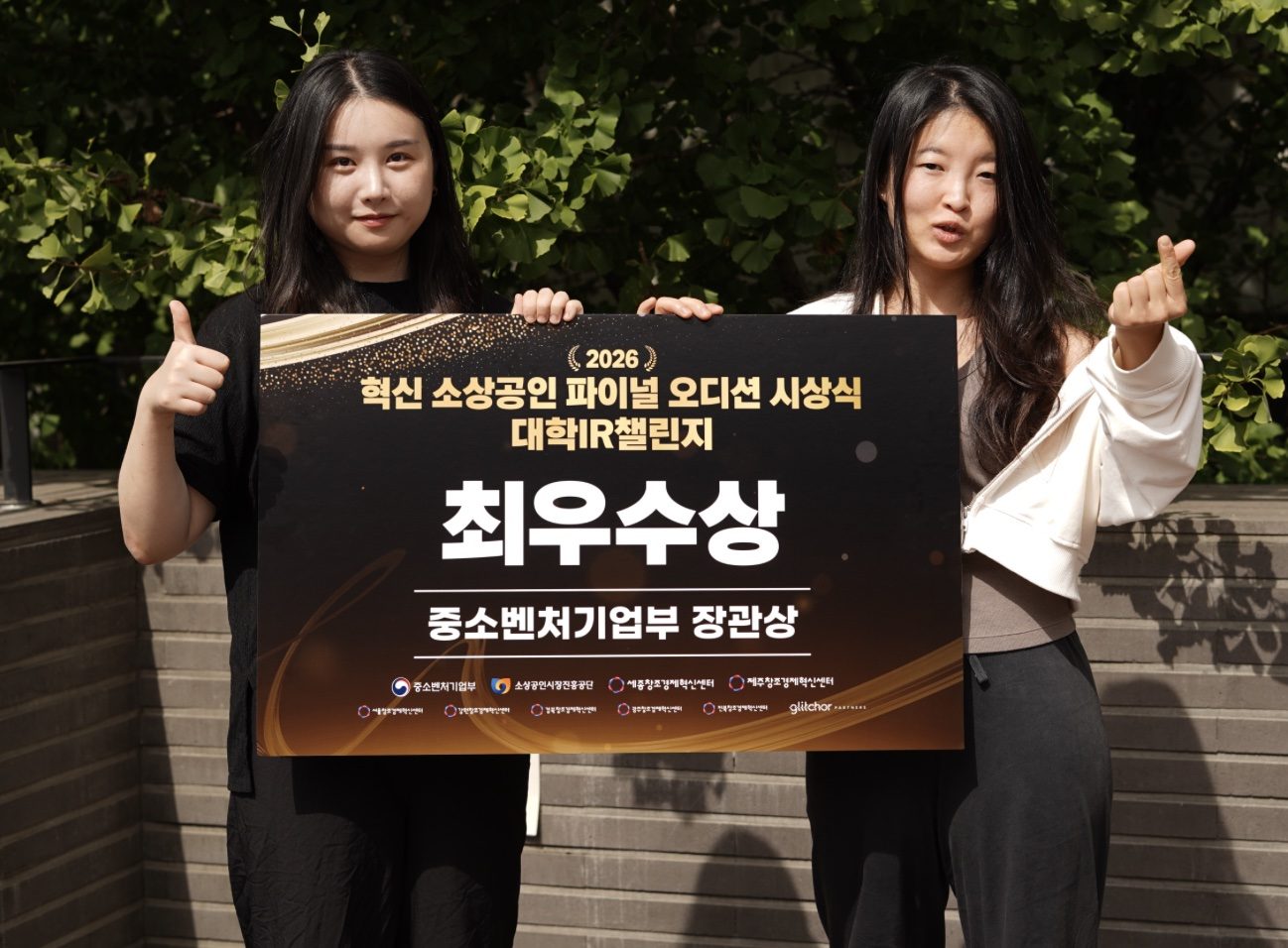 두 공동대표가 나무 앞에서 ‘2026 혁신 소상공인 파이널 오디션 시상식 대학IR챌린지 최우수상 중소벤처기업부 장관상’이라고 적힌 검은 판을 함께 들고 있다.